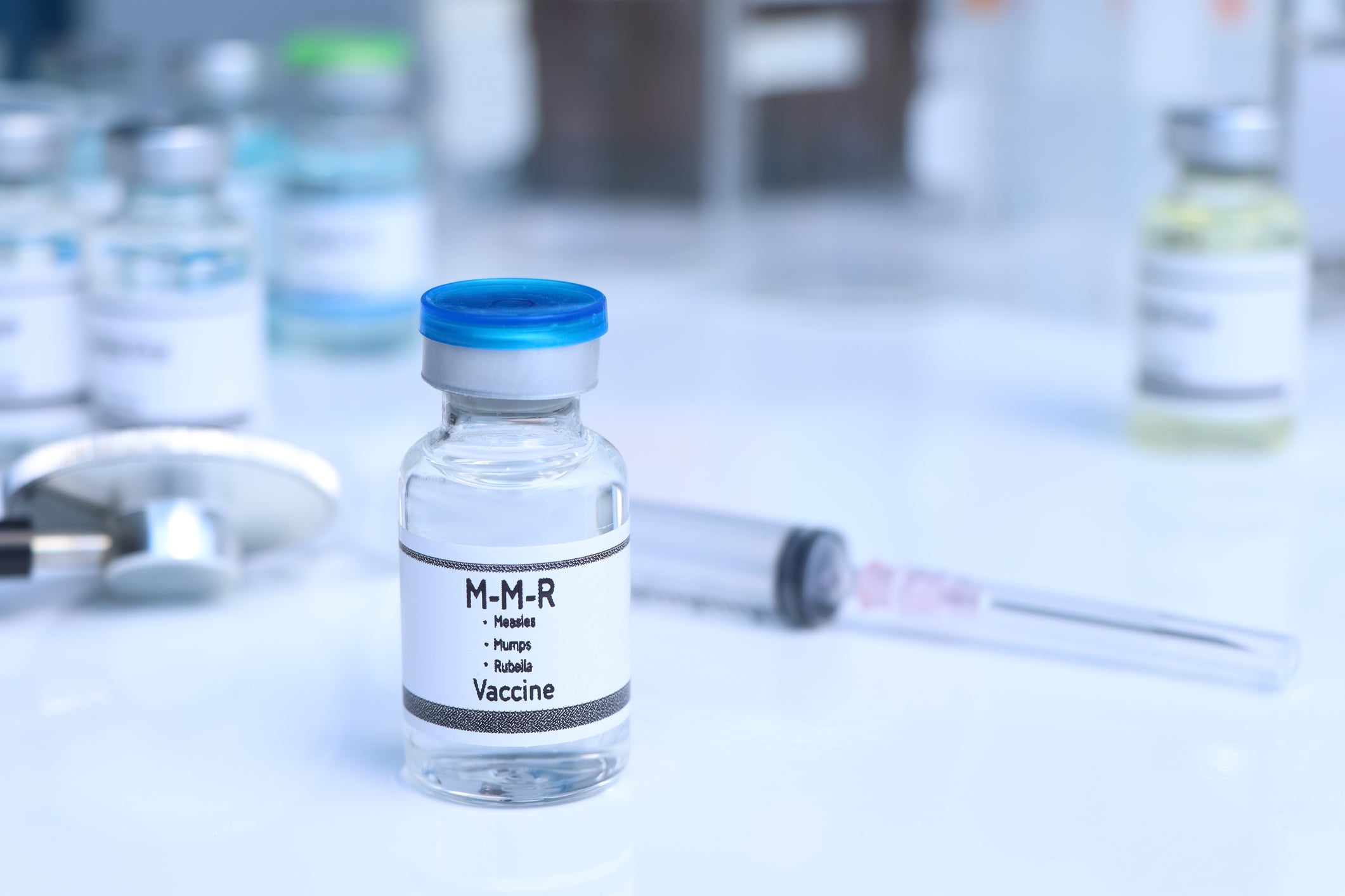 A vial of the measles, mumps, and rubella vaccine sits on a table in front of a syringe and other medical supplies.